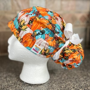Peut inclure: Un bonnet de chirurgien à motifs de récolte d'automne, avec des citrouilles orange, blanches et bleues. Il comporte un nœud blanc et une étiquette indiquant "Mom & Ash & Betty Designs".