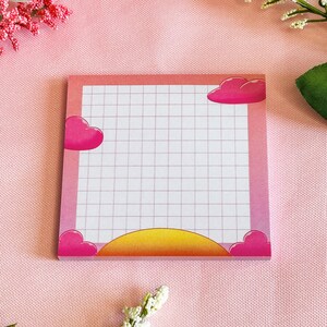 May include: A pink and white grid notepad with a gradient border. The notepad features pink heart and cloud illustrations in each corner and a yellow and orange sunrise design at the bottom. The notepad is designed for taking notes.