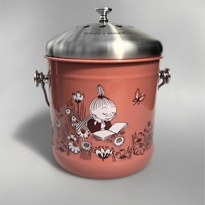Pode incluir: Um caixote de compostagem de metal rosa com uma tampa de aço inoxidável e uma ilustração de um personagem Moomin. O caixote tem uma alça de cada lado e uma pequena abertura na tampa. O texto "BioPots x MOOMIN" está impresso na tampa.