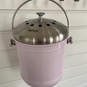 Puede incluir: Un cubo de compost de metal rosa con una tapa plateada y un asa. La tapa tiene un pequeño pomo y agujeros para la ventilación. El cubo tiene el texto "BioProffa" impreso en la tapa.