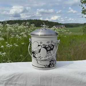 May include: A white and silver compost pail with a lid and handle. The pail features a black and white illustration of cartoon characters and flowers. The background shows a green field and a cloudy sky.