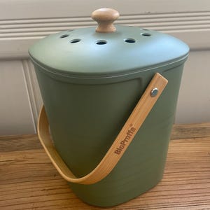 May include: A green compost bin with a wooden handle and lid. The bin has a wooden knob on the lid and the text "BioProffita" printed on the side.