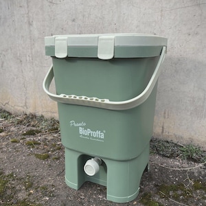 Puede incluir: Un cubo de compostaje verde con asa y tapa blancas. El cubo tiene el texto "Pronto BioProffa" en la parte delantera. Tiene un grifo cerca de la parte inferior y está sobre una superficie de piedra.