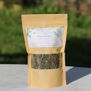 Peut inclure: Un sachet en papier kraft avec une fenêtre transparente, rempli d'herbes séchées. Le sac porte une étiquette avec le texte "Artemisia annua" et des illustrations botaniques. L'arrière-plan est vert flou.
