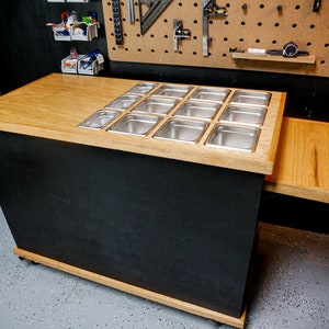 May include: A custom-built prep station in black and wood tones, featuring stainless steel containers. The station has a light wood countertop with a section for the containers and a pull-out side table. It is on wheels.