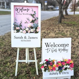 Puede incluir: Dos letreros de boda de madera blanca. Uno tiene un fondo rosa con el texto "Welcome To Our Wedding", los nombres "Sarah & James" y la fecha 02/04/2025. El otro dice "Welcome To Our Wedding" y "Ashely & Chris". Ambos tienen arreglos florales.