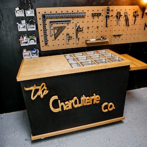 May include: A black and wood charcuterie display with stainless steel food containers. The words "The Charcuterie Co." are displayed in wood lettering on the front. A pegboard with tools hangs in the background.