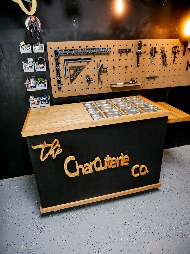 May include: A black and wood charcuterie display with stainless steel food containers. The words "The Charcuterie Co." are displayed in wood lettering on the front. A pegboard with tools hangs in the background.