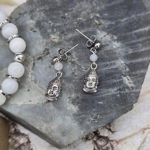 Puede incluir: Un par de pendientes de plata con cuentas blancas y un dije de Buda de plata. Los pendientes están sobre un fondo de piedra gris.