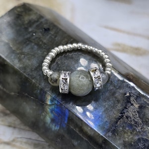 May include: A silver beaded ring with a central gray gemstone and two silver accents with clear stones. The ring is set on a dark, textured surface with blue and green hues.