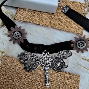 May include: A steampunk-style choker necklace with a black velvet band and a dragonfly pendant. The dragonfly is made of gears and metal components. The necklace also features two gear accents on either side of the band.