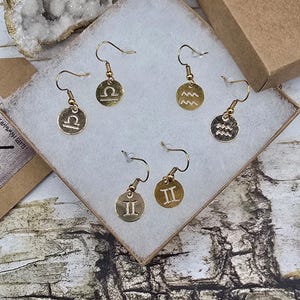 May include: Gold-colored earrings with various zodiac symbols displayed in a white box. The earrings have a hook design and are circular with symbols for Libra, Aquarius, and Gemini. The box is inside a brown cardboard box.