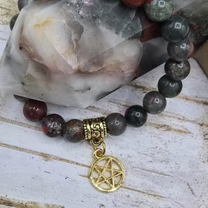 May include: A bracelet made of dark green, red, and grey stones with a gold charm in the shape of a pentagram.