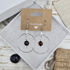 May include: A pair of handmade silver hoop earrings with a small, round, brown and black bead suspended in the center of each hoop. The earrings are displayed in a white box with a brown tag that reads "Handmade" and a heart symbol.