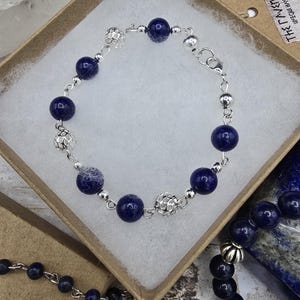 May include: A silver chain bracelet with dark blue lapis lazuli beads and silver spacers. The bracelet is in a white box with a brown paper tag that says "The Gypsy Witch" and "Unique Handmade Gifts".