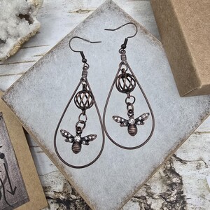 May include: A pair of copper-colored drop earrings. Each earring features a teardrop-shaped frame with a decorative orb and a bee pendant adorned with small, sparkling crystals. The earrings are displayed on a white surface.