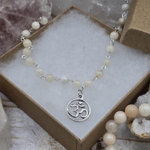 May include: A white beaded necklace with a silver Om charm pendant. The necklace is displayed in a brown box with a white background.