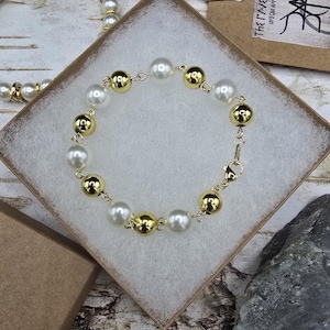 May include: A white and gold pearl bracelet with a clasp closure. The bracelet is displayed in a white box with a brown paper tag that reads "THE GLOWN WEETCH" and "UNIQUE HANDMADE GIFTS".