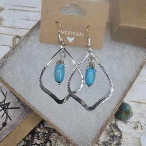 May include: Handmade silver-tone earrings with a teardrop shape. Each earring features a turquoise-colored stone and a small silver accent. The earrings are displayed in a gift box with a 'Handmade' tag.