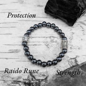 May include: A black hematite beaded bracelet with a silver charm featuring the Raido rune. The bracelet is on a white wooden surface with the words "Protection", "Raido Rune", and "Strength" written below it.