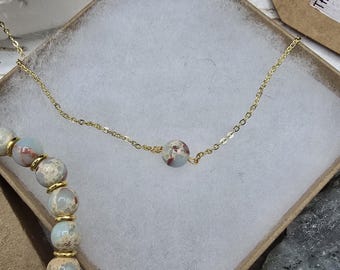 May include: A gold chain necklace with a single light blue and brown gemstone bead. The necklace is displayed in a white box with a light blue and brown beaded bracelet.