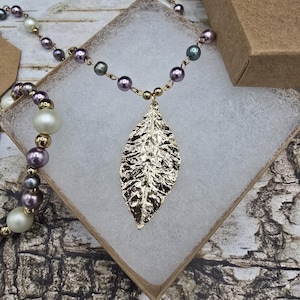 May include: A gold leaf pendant necklace with a beaded chain of white, gold, and purple pearls. The necklace is presented in an open cardboard box with a white lining, and another necklace is visible.