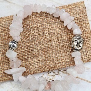 Puede incluir: Una pulsera de cuentas de chips de cuarzo rosa con un dije de cabeza de Buda plateado y una cuenta plateada. La pulsera está hecha de piedras de color rosa claro de forma irregular. La pulsera se exhibe sobre una superficie tejida de color beige.