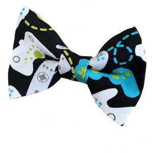 May include: A bow tie featuring a black background with a repeating pattern of white and bright blue video game controllers, accented by small lime green circular details and dashed lines throughout the fabric design of the accessory.