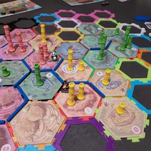 Peut inclure: Un plateau de jeu hexagonal coloré avec différentes pièces en plastique de couleurs, notamment vert, jaune et rose. Il y a aussi de petites pièces en bois avec des visages de panda. Le plateau est en plastique et présente une variété de textures différentes.