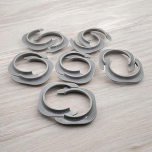 Peut inclure: Six objets gris en forme d'anneau en plastique avec un design incurvé. Chaque anneau a une ouverture fendue. Les objets sont disposés sur une surface en bois de couleur claire. Les anneaux semblent être des composants d'interverrouillage ou de connexion.