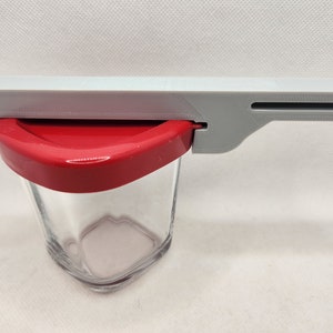 Pode incluir: Um abridor de frascos de plástico cinza com uma tampa vermelha, projetado para abrir frascos com um selo hermético.