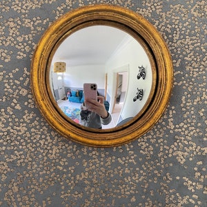 Pu&ograve; includere: Uno specchio rotondo con cornice dorata e lente convessa, che riflette una persona che scatta un selfie con un telefono rosa. Lo specchio &egrave; appeso a una parete con un motivo a macchie grigie e dorate.