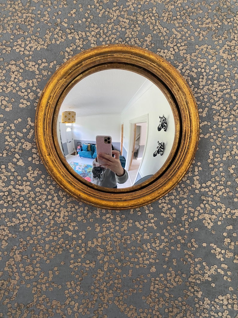 Pu&ograve; includere: Uno specchio rotondo con cornice dorata e lente convessa, che riflette una persona che scatta un selfie con un telefono rosa. Lo specchio &egrave; appeso a una parete con un motivo a macchie grigie e dorate.