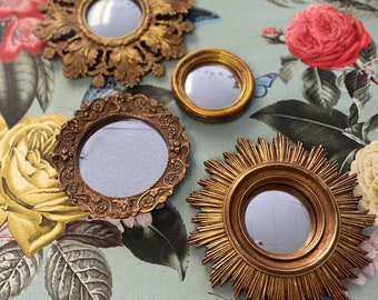 Può includere: Una collezione di piccoli specchi dorati con varie cornici decorative. Gli specchi, di forme diverse tra cui rotonde e a forma di sole, sono disposti su una superficie a motivi floreali, mostrando i loro design ornati.