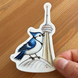 Puede incluir: Una pegatina troquelada que muestra un arrendajo azul posado sobre una superficie curva junto a una torre alta y esbelta con una aguja puntiaguda. El pájaro tiene plumas azules y blancas, y la torre está representada en tonos beige, gris y blanco.