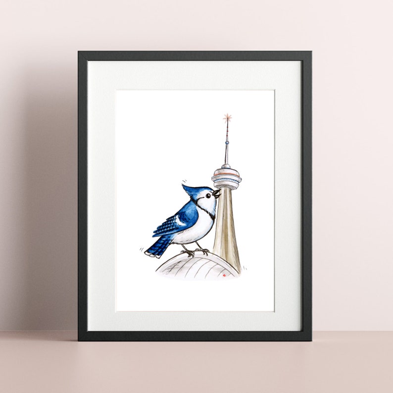 May include: A framed art print featuring a blue jay perched on the CN Tower in Toronto. The bird is depicted with detailed blue and white feathers, set against the tower's beige and white structure. The artwork is framed in a black border.