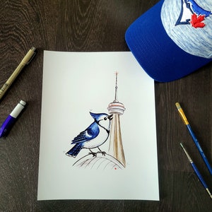 May include: A blue jay illustration perched on a brown structure, possibly a tower, with a blue and white baseball cap featuring a bird and maple leaf logo. The artwork is on white paper, with art supplies nearby.
