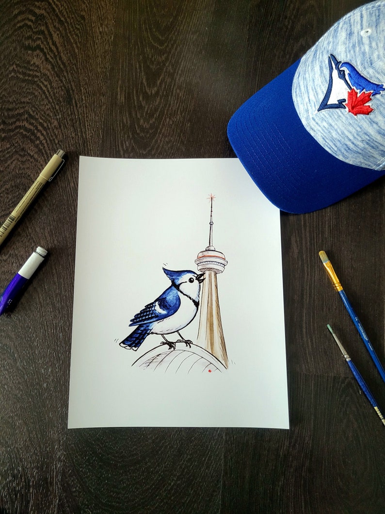 May include: A blue jay illustration perched on a brown structure, possibly a tower, with a blue and white baseball cap featuring a bird and maple leaf logo. The artwork is on white paper, with art supplies nearby.