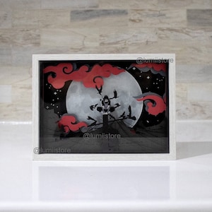 May include: A black and white papercraft shadow box featuring a silhouette of a person standing on a rooftop with crows flying around them. The background is a dark blue sky with a large, full moon and red clouds.