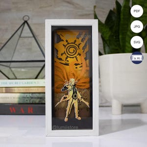 May include: A white framed 3D papercraft of Naruto Uzumaki in his Nine-Tailed Fox form. The papercraft is illuminated from behind, creating a glowing effect. The image is framed in a white frame with a black mount.