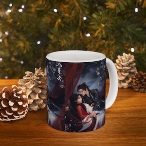 May include: White ceramic mug featuring a romantic scene with two people in traditional clothing. The mug has a wraparound design with a dark background and red accents. The mug is on a wooden surface with pine cones.