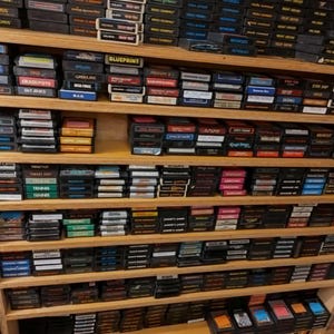 May include: A wall shelf filled with rows of vintage video game cartridges. The cartridges are organized by color and title. The shelf is made of wood and has a light brown finish.