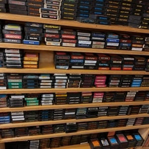 May include: A wooden shelf unit filled with rows of vintage video game cartridges. The cartridges are organized by color and title. The shelf unit is made of wood and has a light brown finish.