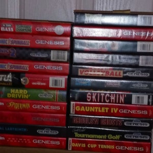 May include: A stack of Sega Genesis video game cartridges. Titles include "Sonic Spinball", "Spiderman", "Fatal Labyrinth", and "Davis Cup Tennis Genesis". The cartridges are primarily red and black, with white and yellow text.