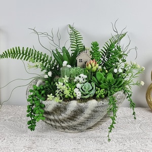 Puede incluir: Un arreglo decorativo de suculentas en una maceta de cerámica blanca con un acabado rústico. La maceta tiene forma de concha y presenta una pequeña figura de casa. El arreglo incluye varios tipos de suculentas, helechos y flores blancas.