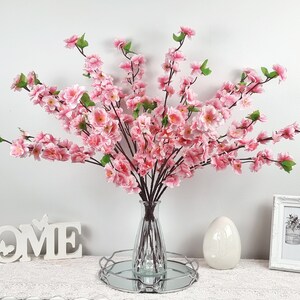May include: A bouquet of pink artificial cherry blossom branches in a clear glass vase on a silver tray. The vase is on a white surface with a white wooden sign that says "HOME" and a white picture frame.