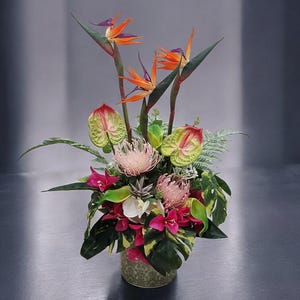 Può includere: Una vivace composizione floreale con fiori di uccello del paradiso con fiori arancioni e viola, completata da protee rosa e anthurium. La composizione è in un vaso verde decorativo, circondato da un rigoglioso fogliame verde.