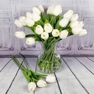 May include: A bouquet of white artificial tulips in a clear glass vase. The vase is sitting on a white wooden surface. There are extra tulips on the surface.