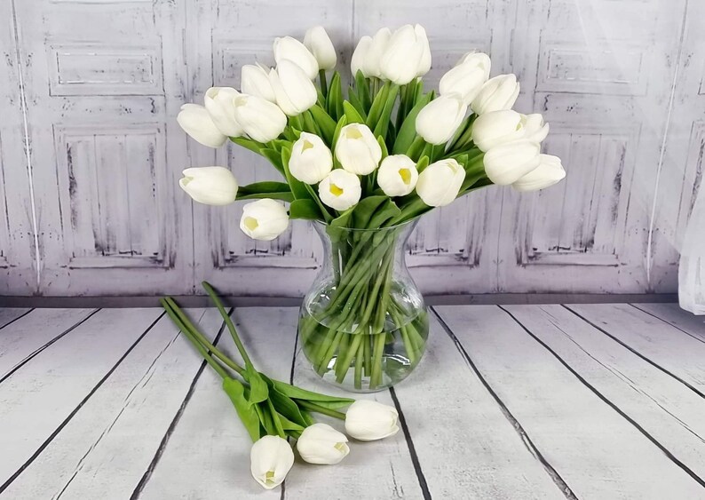 May include: A bouquet of white artificial tulips in a clear glass vase. The vase is sitting on a white wooden surface. There are extra tulips on the surface.