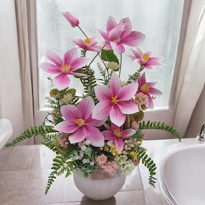 May include: A white ceramic vase with pink and white artificial flowers. The vase is sitting on a white tiled surface.